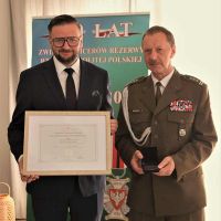 Obok mężczyzny w garniturze trzymającego obraz w ramce stoi mężczyzna w mundurze wojskowym.