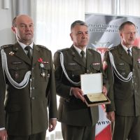 Trzech mężczyzn w mundurach wojskowych stojących obok siebie. Środkowy trzyma w rękach ryngraf w pudełku.