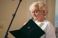 Kobieta trzymająca w lewej ręce otwartą teczkę, a w prawej mikrofon.