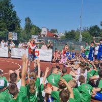 Mężczyzna w stroju sportowym mówi z grupą młodzieży w strojach sportowych siedzących na boisku do koszykówki.