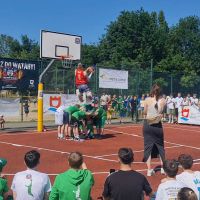 Mężczyzna z piłką do koszykówki w wyskoku nad grupą sześciu chłopców pochylonych do siebie.