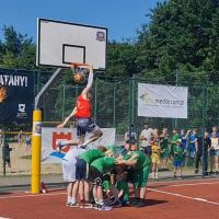 Mężczyzna wkładający piłkę do kosza w wyskoku nad grupą sześciu chłopców pochylonych do siebie.