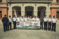 Kobieta wśród kilkunastu mężczyzn w mundurach Marynarki Wojennej stojących za banerem z okrętem wojennym.