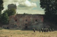 Kilkunastu mężczyzn biegnących z bronią w mundurach wojskowych z I wojny światowej w stronę zabytkowego budynku.