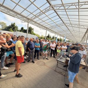 Dwie kobiety i kilkudziesięciu mężczyzn stojących przed dwoma klatkami z gołębiami.