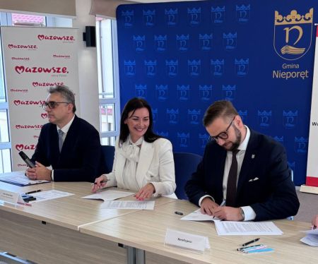 burmistrz podpisujący umowę, obok uśmiecha sie kobieta oraz mężczyzna z mikrofonem