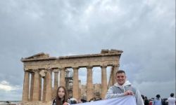 dwie osoby trzymają białą flagę z czarnymi napisami, w tle ruiny Akropolu,