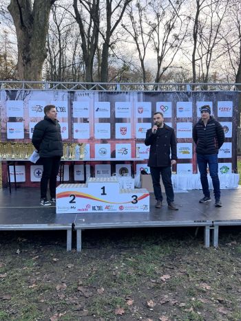 trzy osoby przy podium, zastępca burmistrza trzyma mikrofon, wita zawodników