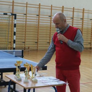 Mężczyzna z mikrofonem w ręku stojący przy stoliku z dyplomami i pucharami.