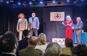 scena, dwóch mężczyzn na scenie, w tle wisi flaga z czerwonym krzyżem napisem pck