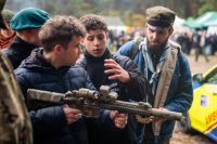 Trzech mężczyzn stojących obok siebie, jeden z nich trzyma w dłoniach karabin.