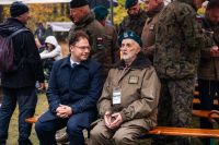 Mężczyzna zwrócony twarzą do siedzącego obok niego starszego mężczyzny w berecie i mundurze wojskowym.