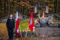 Harcerka stojąca obok flag oraz pomnika ze zniczami i wiązankami kwiatów.