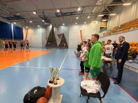 Puchary i nagrody dla zawodników obok mężczyzna w stroju sportowym i mikrofonem oraz mężczyzna w garniturze.