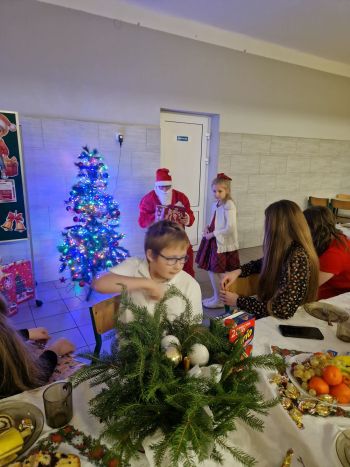 dziewczynka stoi przy Mikołaju, dzieci siedzące przy stole wigilijnym