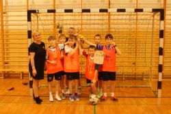 Siedmiu chłopców w strojach sportowych z medalami na szyjach trzymają wspólnie puchar uniesiony do góry.