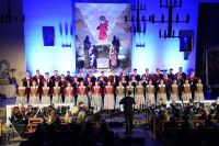 Śpiewające w kościele kobiety i mężczyźni ubrani w różnokolorowe stroje ludowe.