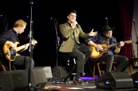 Mężczyzna z mikrofonem siedzący pomiędzy dwoma mężczyznami grającymi na gitarach.