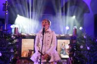 Kobieta śpiewająca do mikrofonu, stojąca na tle szopki bożonarodzeniowej.