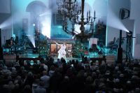 Kilkadziesiąt kobiet i mężczyzn stojących przed kobietą śpiewającą do mikrofonu, stojącą na tle szopki bożonarodzeniowej.