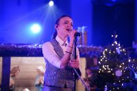 Kobieta śpiewająca do mikrofonu, stojąca na tle szopki bożonarodzeniowej.