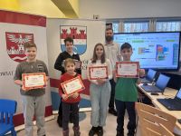 Dzieci stojące z dyplomami w rękach. Za nimi mężczyzna, dwa rollupy i duży monitor.