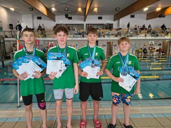 Czterech chłopców w strojach sportowych z medalami na szyjach i dyplomami w rękach, za nimi basen