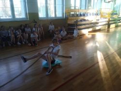 Dziewczynki w strojach sportowych na sali gimnastycznej, jedna z nich ciągnie linę siedząc na wózku transportowym.