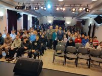 Siedząca na krzesłach młodzież i nauczyciele, w środku stoi kobiet i trzech mężczyzn, dwóch z nich w mundurach wojskowych.