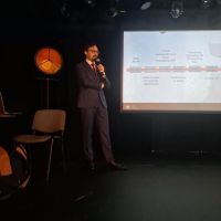 Mężczyzna trzymający mikrofon w dłoni, po prawej stronie ekran z wyświetlonym slajdem z napisami i grafiką.