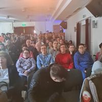 Młodzież i dorośli siedzący w sali na krzesłach.