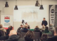 Mężczyzna mówiący do grupy osób, za nim wyświetlony slajd na ścianie, obok roll-up.