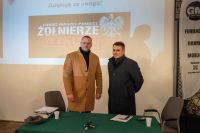 Dwóch mężczyzn stojących za stołem, za nimi wyświetlony slajd na ścianie, obok flaga reklamowa.