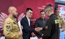 zgromadzona młodzież w strojach czarnych strojach pożarniczych, wręczanie nagród, w tle czerwona ścianka straży pożarnej