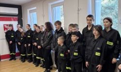 zgromadzona młodzież w strojach czarnych strojach pożarniczych, wręczanie nagród, w tle czerwona ścianka straży pożarnej