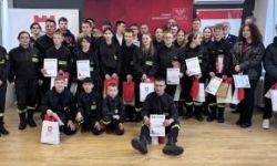 zgromadzona młodzież w strojach czarnych strojach pożarniczych, wręczanie nagród, w tle czerwona ścianka straży pożarnej