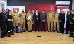 zgromadzona młodzież w strojach czarnych strojach pożarniczych, wręczanie nagród, w tle czerwona ścianka straży pożarnej