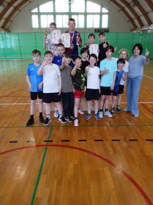 Grupa chłopców stojących na parkiecie i podium, trzech z medalami, pucharami i dyplomami, obok kobieta.