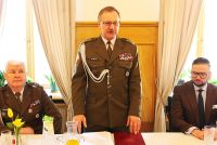 Stojący przy stole żołnie obok siedzący żołnierz i mężczyzna w garniturze.