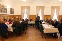 Kobiety i mężczyźni w mundurach siedzący za stołami, jeden mężczyzna w garniturze stoi za stołem.