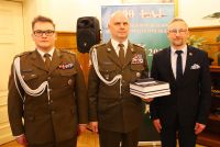 Trzech stojących mężczyzn: dwóch w mundurach wojskowych, jeden z nich trzyma książki w ręku, obok mężczyzna w garniturze.