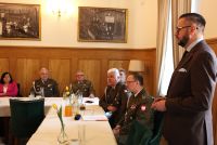 Mężczyzna w garniturze, stojący za stołem mówiący do kobiet i żołnierzy siedzących za stołami.