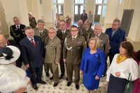 Kobiety i mężczyźni w garniturach i mundurach wojskowych stojący w holu zabytkowego budynku.