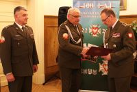 Dwóch mężczyzn w mundurach wojskowych ściska sobie prawe dłonie, jeden z nich trzyma w lewej ręce otwartą teczkę.