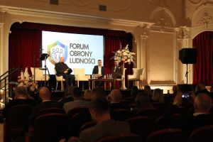 Trzech mężczyzn siedzących w fotelach na scenie, za nimi slajd na ekranie, poniżej grupa osób siedzących w fotelach kinowych.