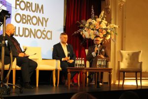 Trzech mężczyzn w garniturach, siedzących w fotelach, jeden z nich mówiący do mikrofonu.