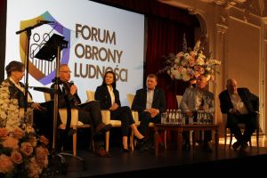 Dwie kobiet i czterech mężczyzn siedzących w fotelach, za nimi slajd na ekranie, jeden z nich mówiący do mikrofonu.