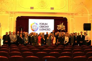 Uczestnicy Forum stojący obok siebie, za nimi slajd Forum Obrony Ludności.