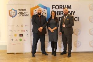 Kobieta stojąca pomiędzy dwoma mężczyznami, za nimi roll-upy Forum Obrony Ludności, poniżej logotypy.