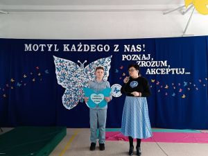 Obok kobiety mówiącej do mikrofonu stoi chłopiec trzymający serce w dłoniach.